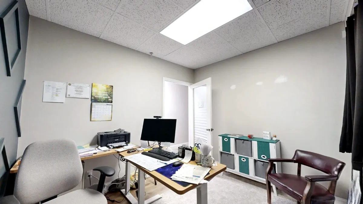 Office 211 desk with monitor and printer, gray task chair, burgundy guest chair, and teal-and-gray storage bins.