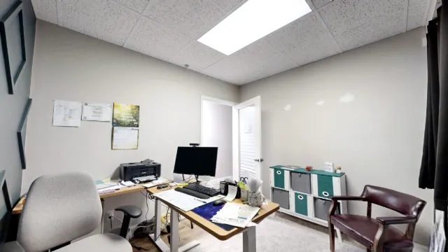 Office 211 desk with monitor and printer, gray task chair, burgundy guest chair, and teal-and-gray storage bins.