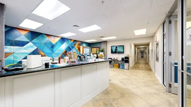 Brochure-lined reception counter beside glass entry doors, with a wall-mounted television and long corridor beyond.