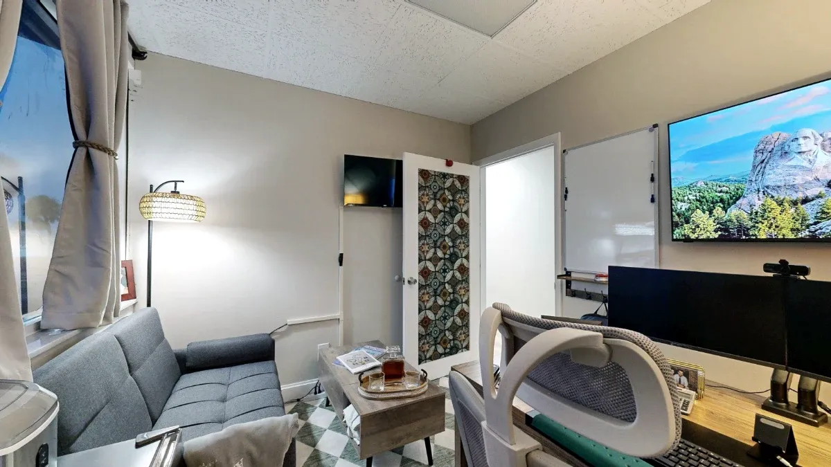 Reverse office view shows desk monitors, a mesh chair, whiteboard and wall screens opposite the sofa and coffee table.