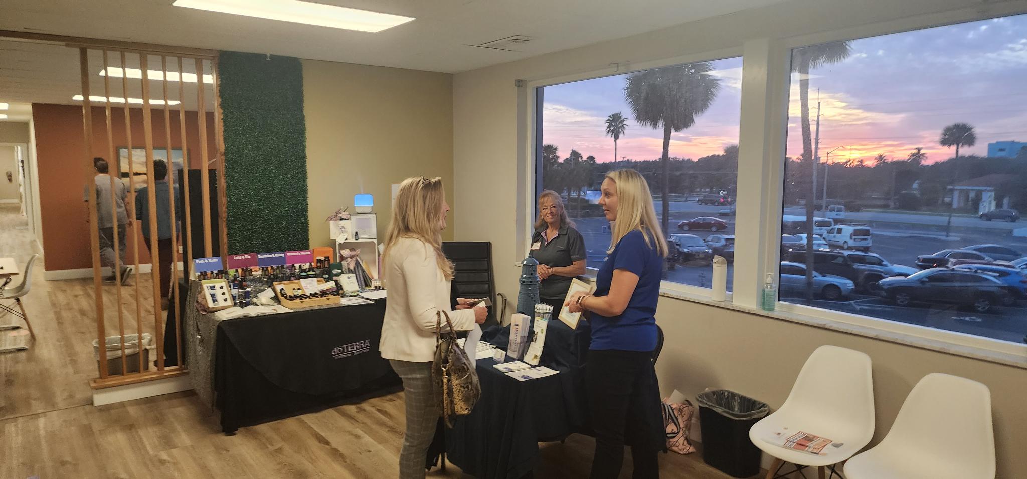 Three women chat beside product display tables, with palm trees and a sunset visible through large windows.