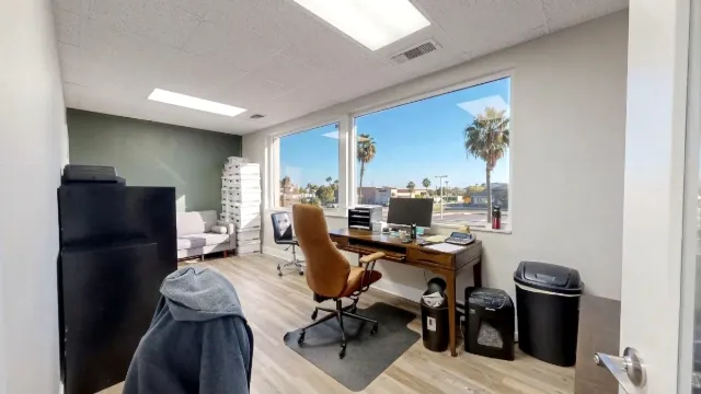 A wooden desk and tan high-back chair face broad windows overlooking palm trees, with a sofa and storage at the back.