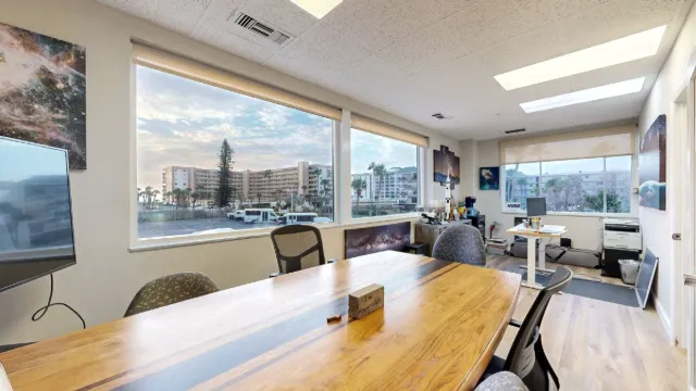 Wide view across office 203's meeting table toward a rear workstation, showing all three windows and a printer.