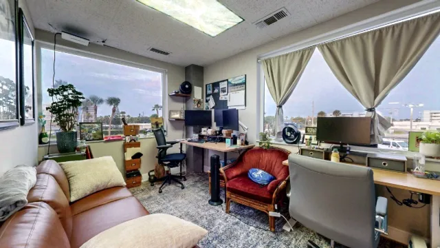 Private office with two desks, monitors, large curtained windows, a brown sofa, a red armchair and potted plants.