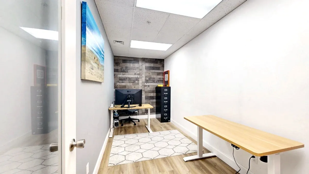 Doorway view of a narrow office with two wood-top desks, a patterned rug, a black filing cabinet and a wood-look accent wall.