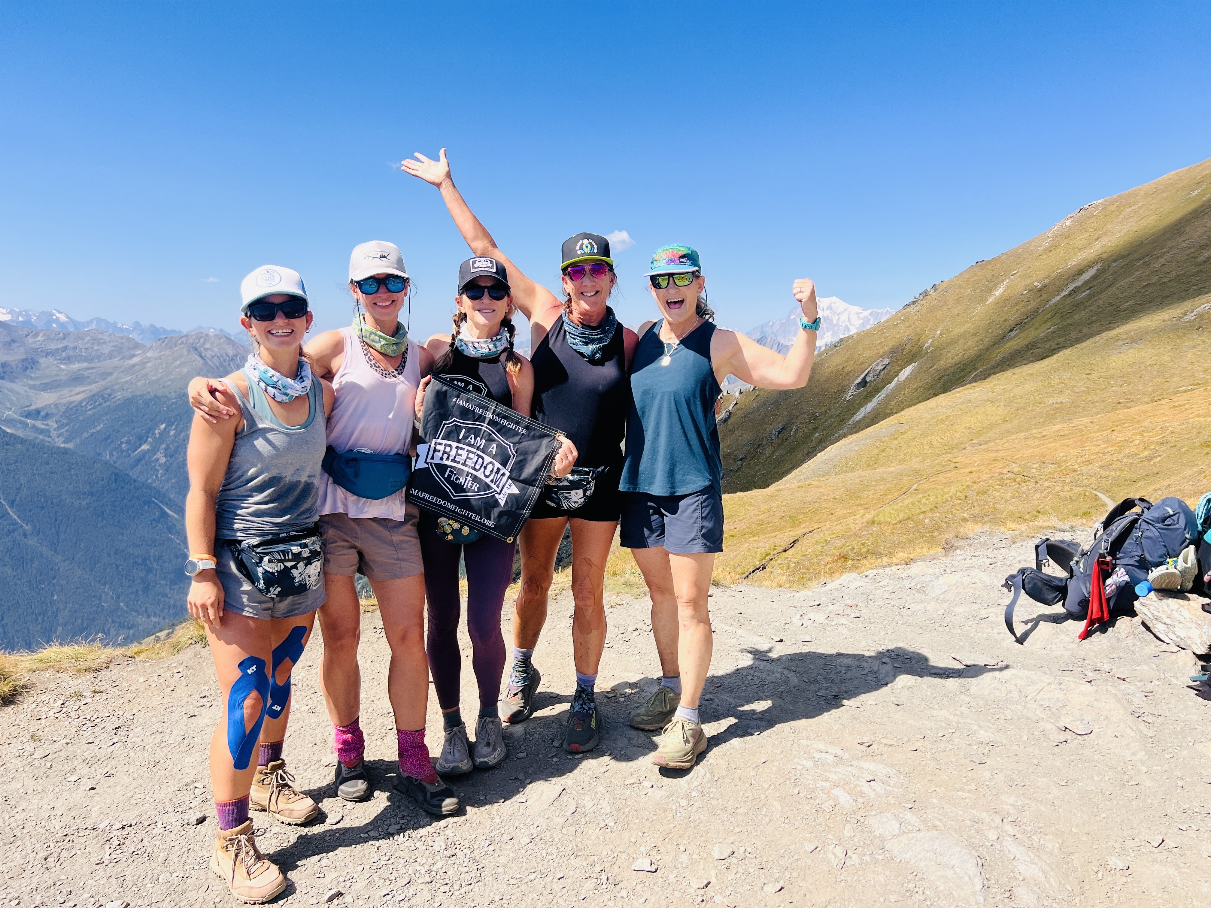 Five hikers pose on a mountain trail with an “I Am a Freedom Fighter” banner and distant snowy peaks behind them.