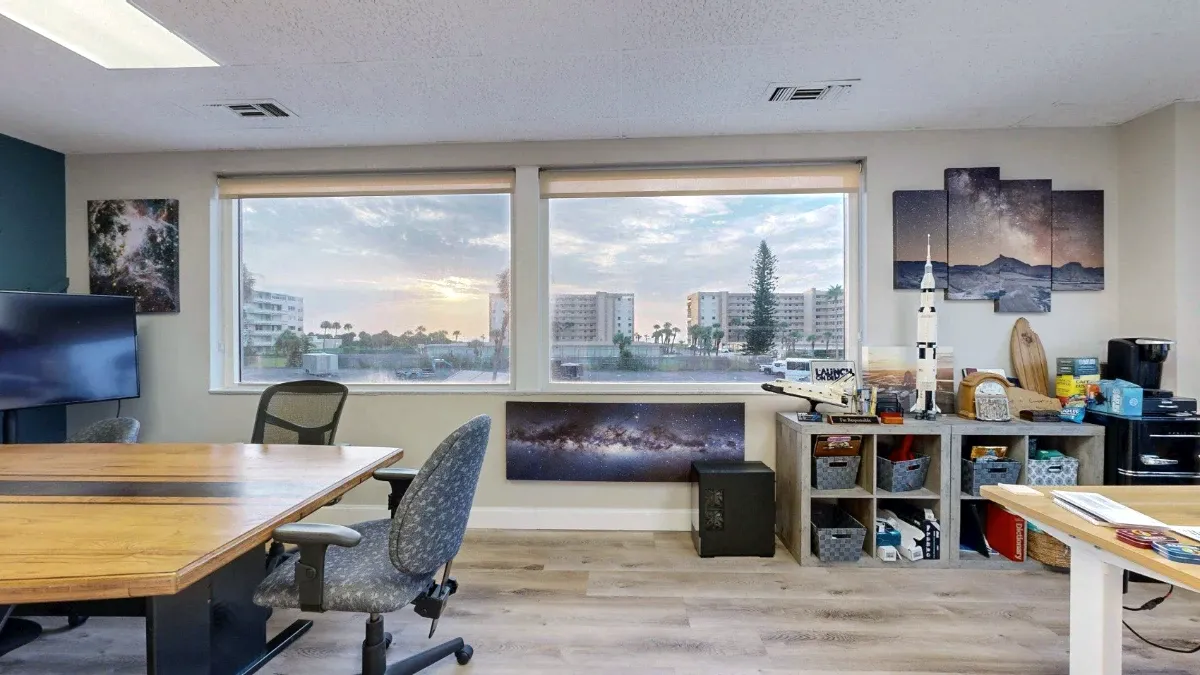 Two large windows above galaxy artwork in office 203, with meeting chairs at left and cubby shelves with models at right.