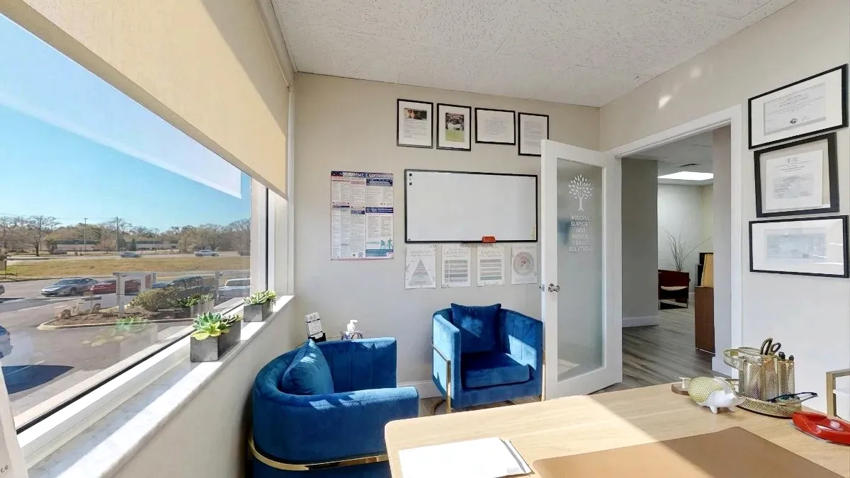 Small office with a large window, two blue chairs, a wood desk, a whiteboard and an open frosted-glass door.