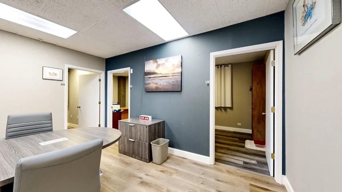 Gray meeting chairs and a table face a blue accent wall with beach artwork, a filing cabinet and open office doorways.