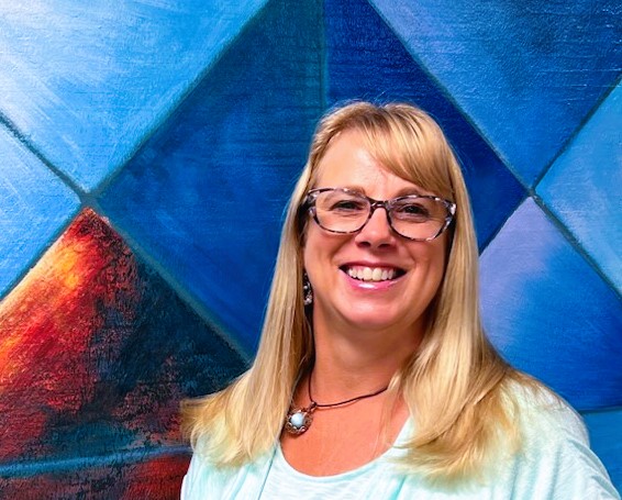 A smiling blonde woman wearing glasses and a light blue top stands in front of a colorful geometric wall.