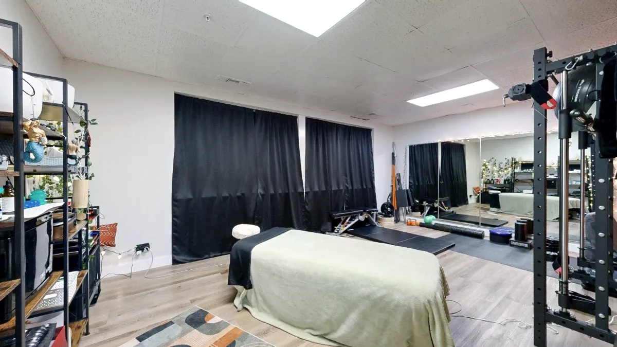 Massage table in Office 227 with two black-curtained windows, supply shelves and a mirrored exercise area behind it.