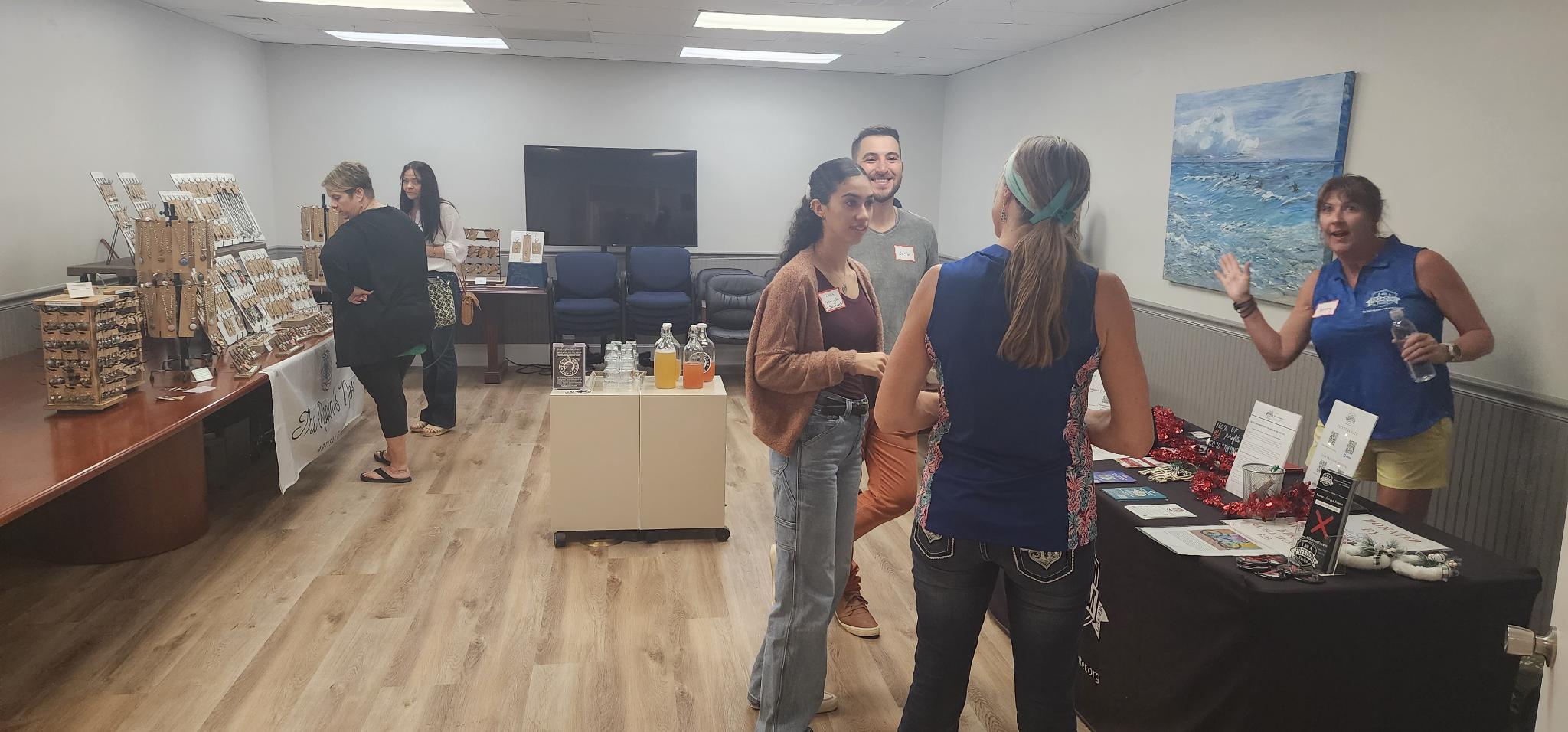People chat and browse jewelry displays in a room with an information table, bottled drinks, chairs, and a television.