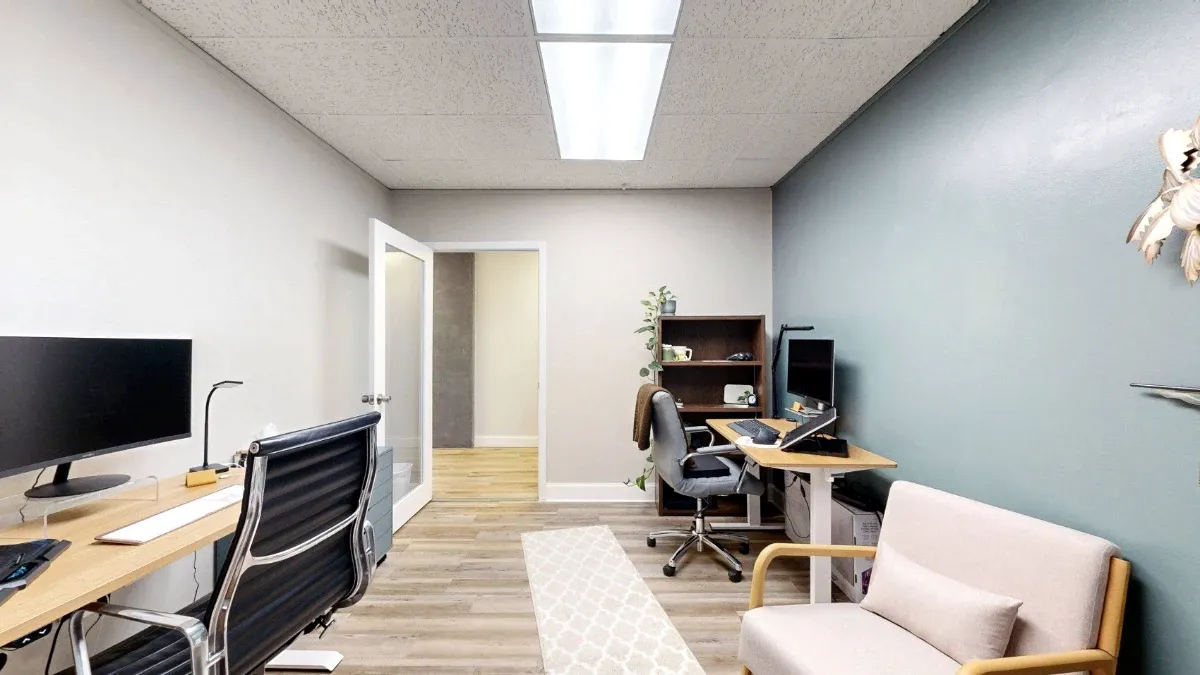 View toward Office 222A's open doorway, showing two desks, a bookcase and a cushioned armchair against the teal wall.