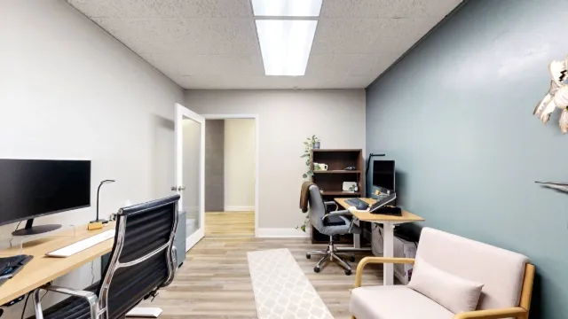 View toward Office 222A's open doorway, showing two desks, a bookcase and a cushioned armchair against the teal wall.
