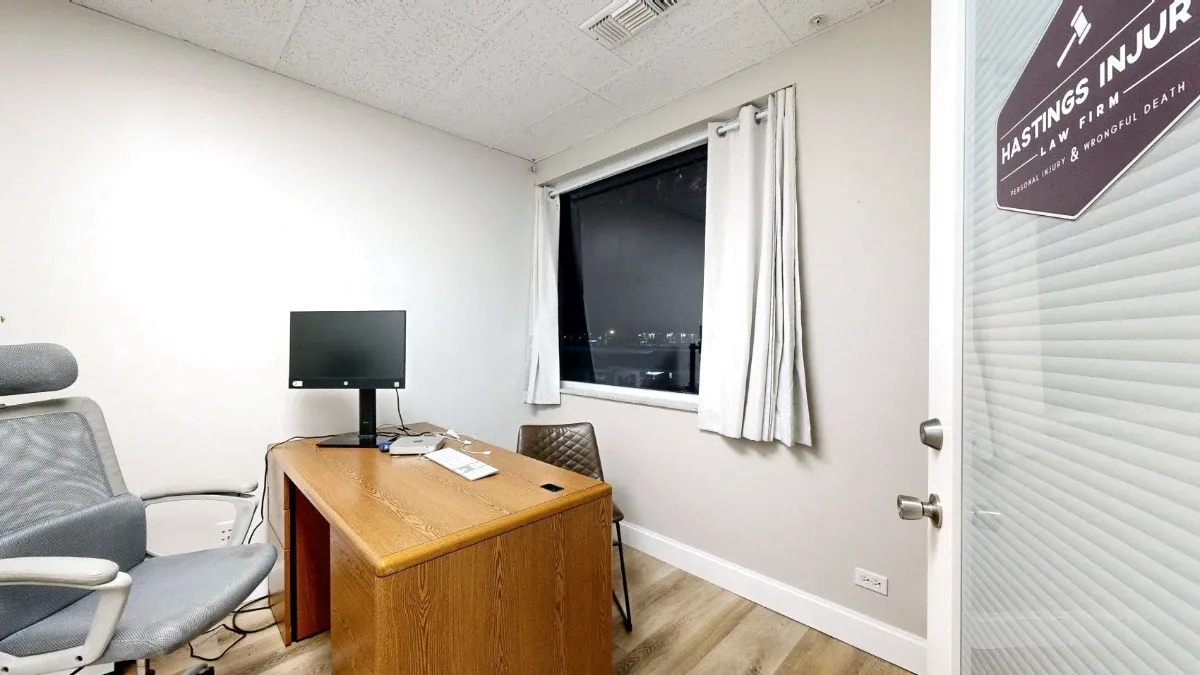 Office 221 contains a wooden desk, gray office chair and brown guest chair beside a north-facing window with white curtains.