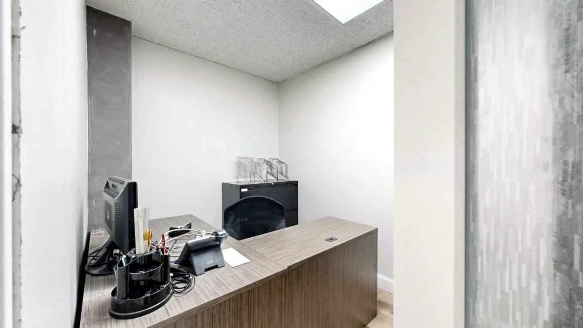 Small office with an L-shaped wood-look desk, monitor, phone, black chair, and filing cabinet beneath overhead lighting.