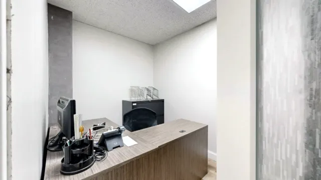 Small office with an L-shaped wood-look desk, monitor, phone, black chair, and filing cabinet beneath overhead lighting.