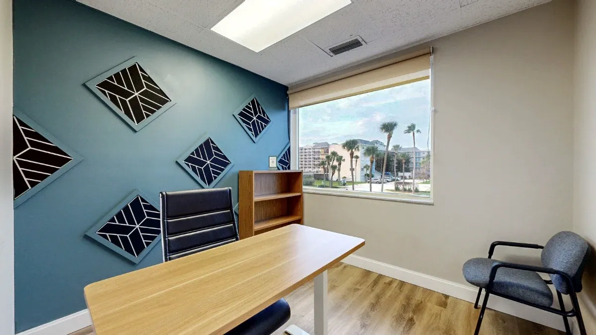A desk and black office chair face a teal wall with geometric artwork, beside a bookcase, window and gray guest chair.