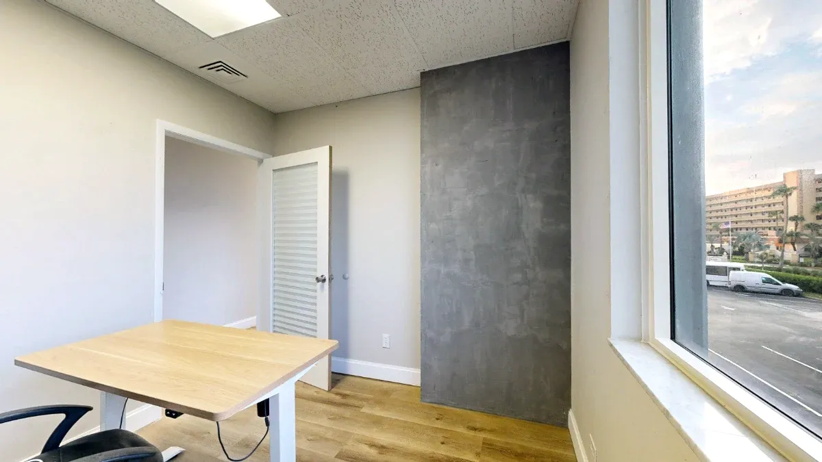 Office 204 viewed beside the window, with a wood-top desk, tall gray wall panel and open door.