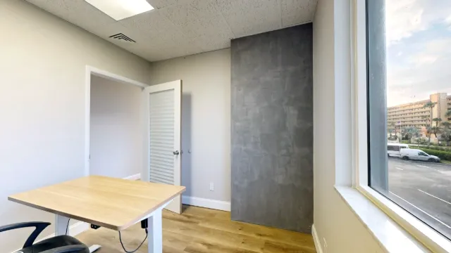 Office 204 viewed beside the window, with a wood-top desk, tall gray wall panel and open door.