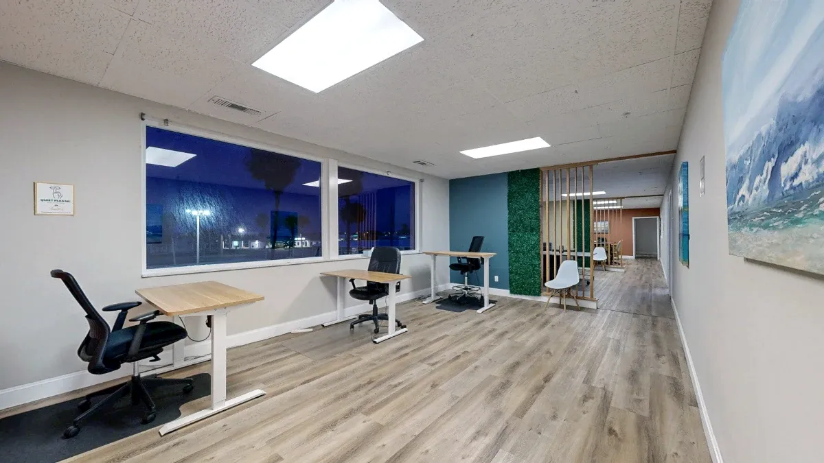 Three desks line dark windows, with ocean artwork on the right and a white chair beside a slatted divider.