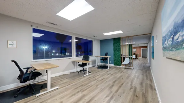 Three desks line dark windows, with ocean artwork on the right and a white chair beside a slatted divider.