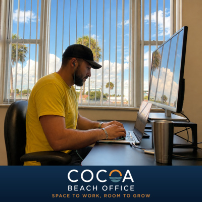 A person in a yellow shirt works on a laptop beside a large monitor and windows, above a Cocoa Beach Office branding strip.