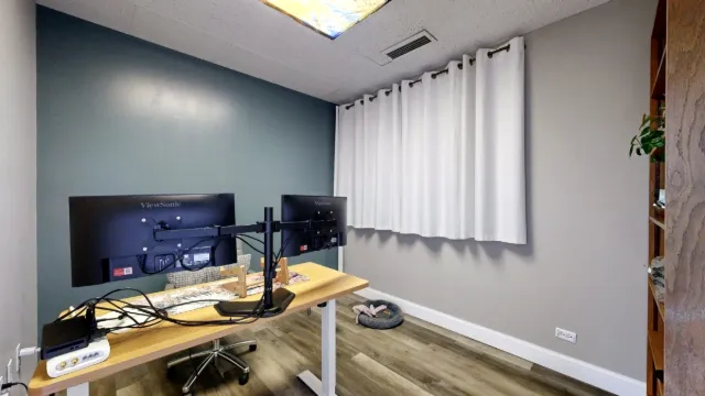 Small office with the backs of two desk monitors, a blue accent wall, white window curtains and wood-look flooring.