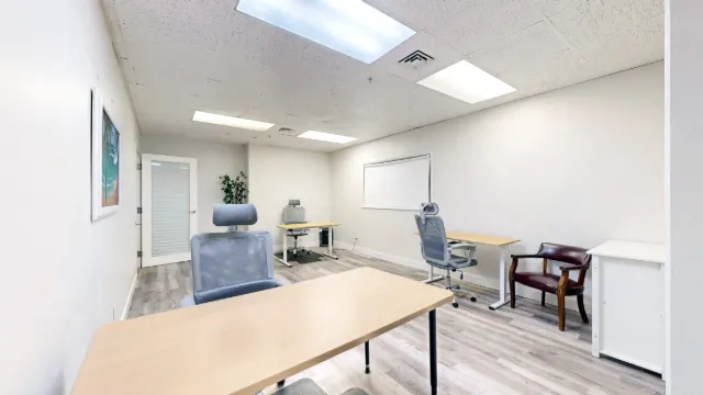View across a foreground desk toward Office 229's entry door, with mesh chairs, a guest chair and a white cabinet.