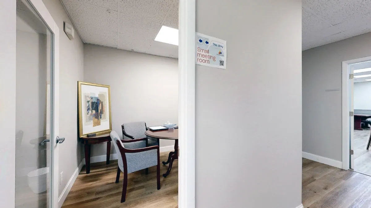 Hallway view into The Cape, with a small meeting room sign, gray chairs, a round table and framed artwork.