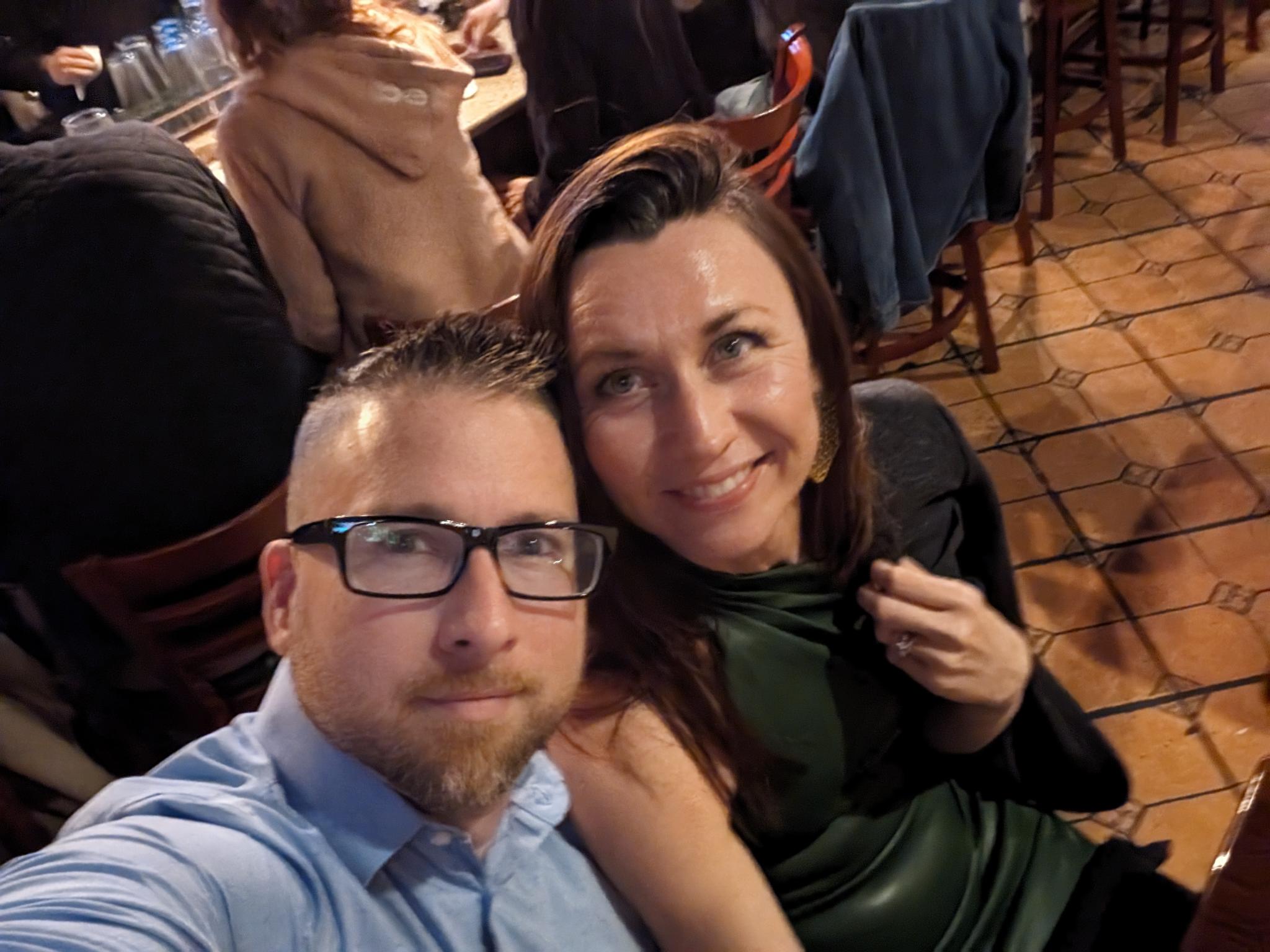 Two adults pose for a selfie with seated patrons, wooden chairs and a terracotta-tiled floor behind them.