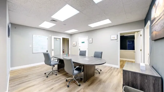 A rounded meeting table with four gray rolling chairs stands between a wall-mounted whiteboard and surrounding office doors.