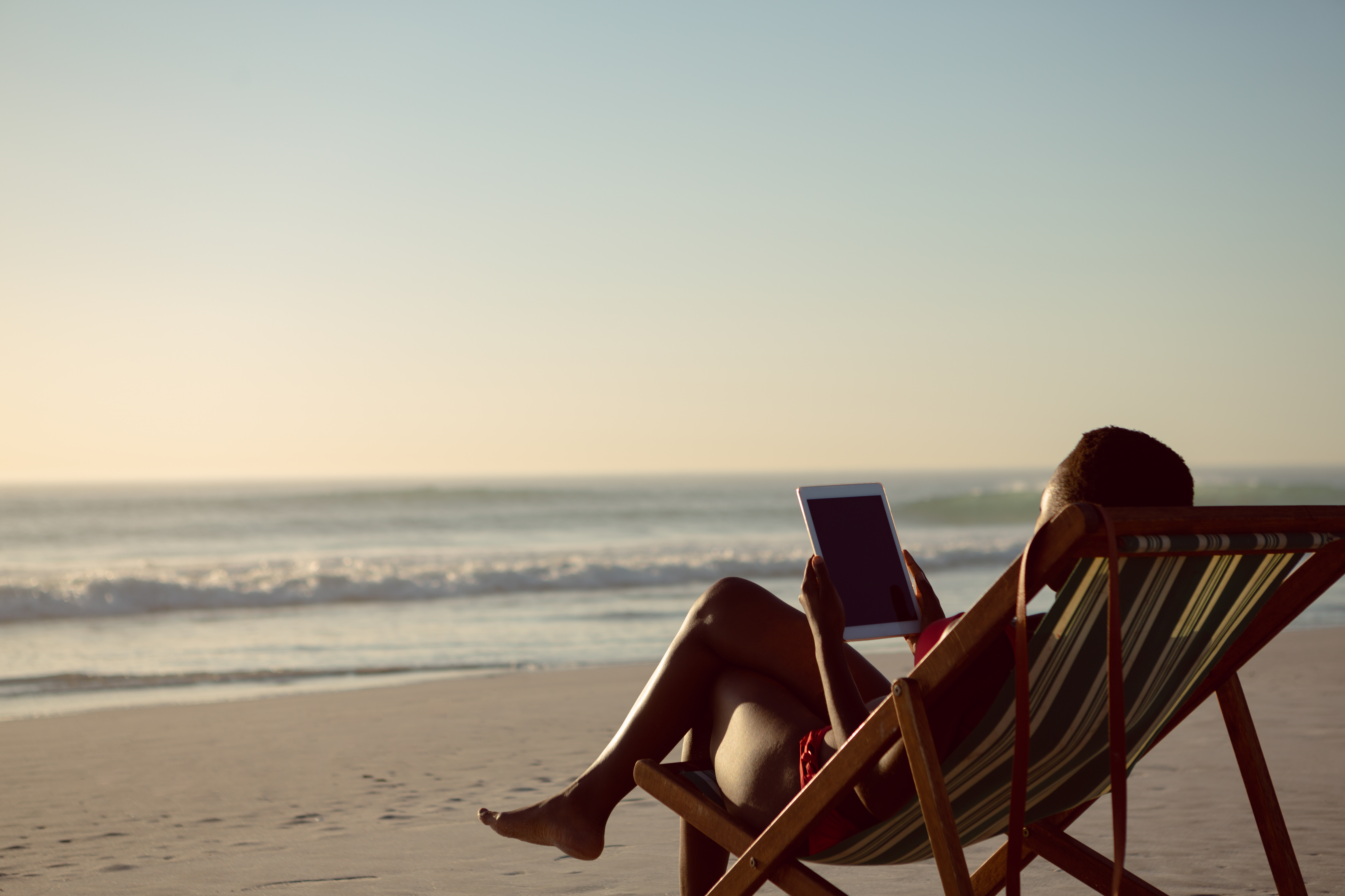 A person reclines in a striped wooden beach chair holding a tablet, with ocean waves in the background.