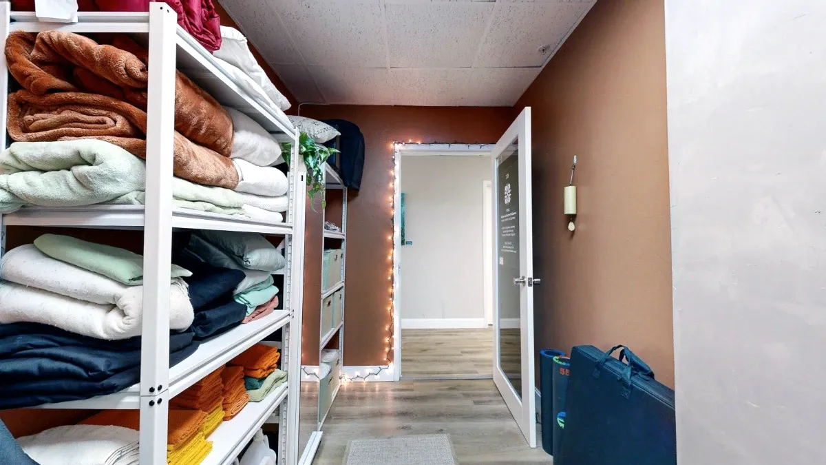 View toward Office 227's open exit door, with folded linens on white shelves and blue equipment cases along the wall.