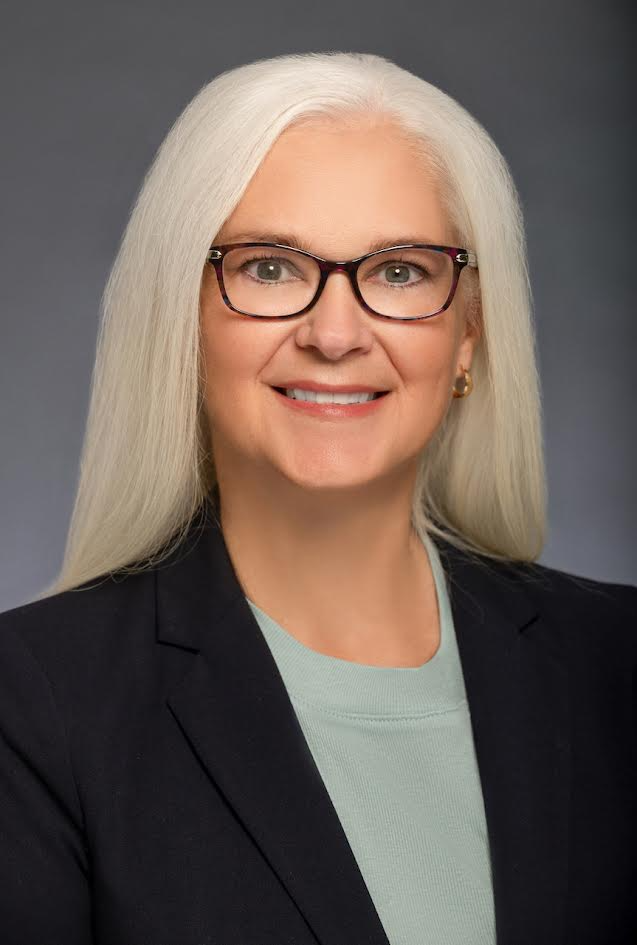 A smiling woman with long white hair and glasses wears a black blazer over a pale green top against a gray background.