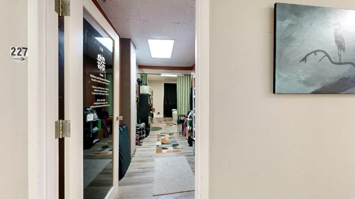 Open glass door into Office 227, with a directional number sign, hallway bird artwork and rugs leading inside.