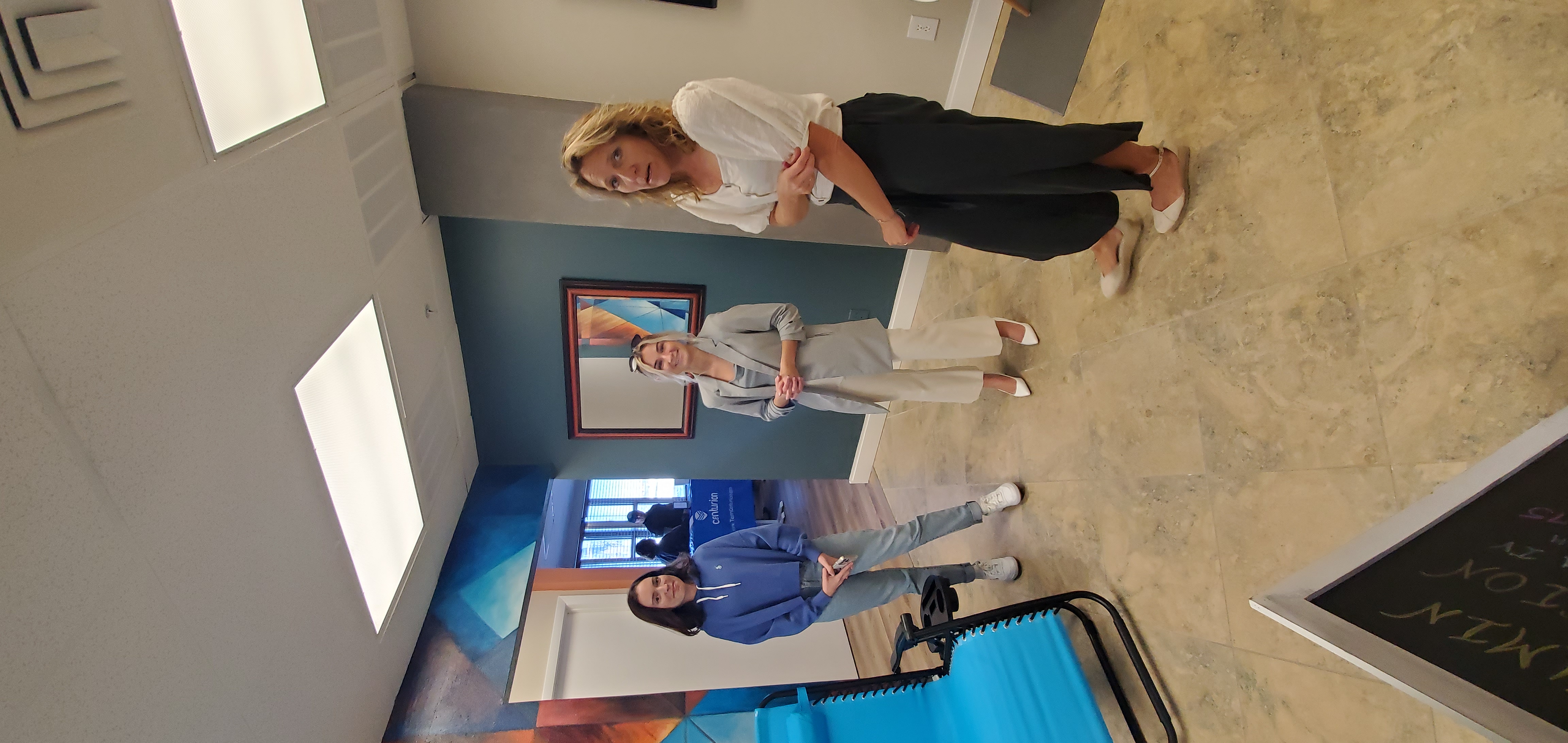 Three people stand beside a turquoise lounge chair, with colorful walls, a framed mirror and a Centurion table in the background.