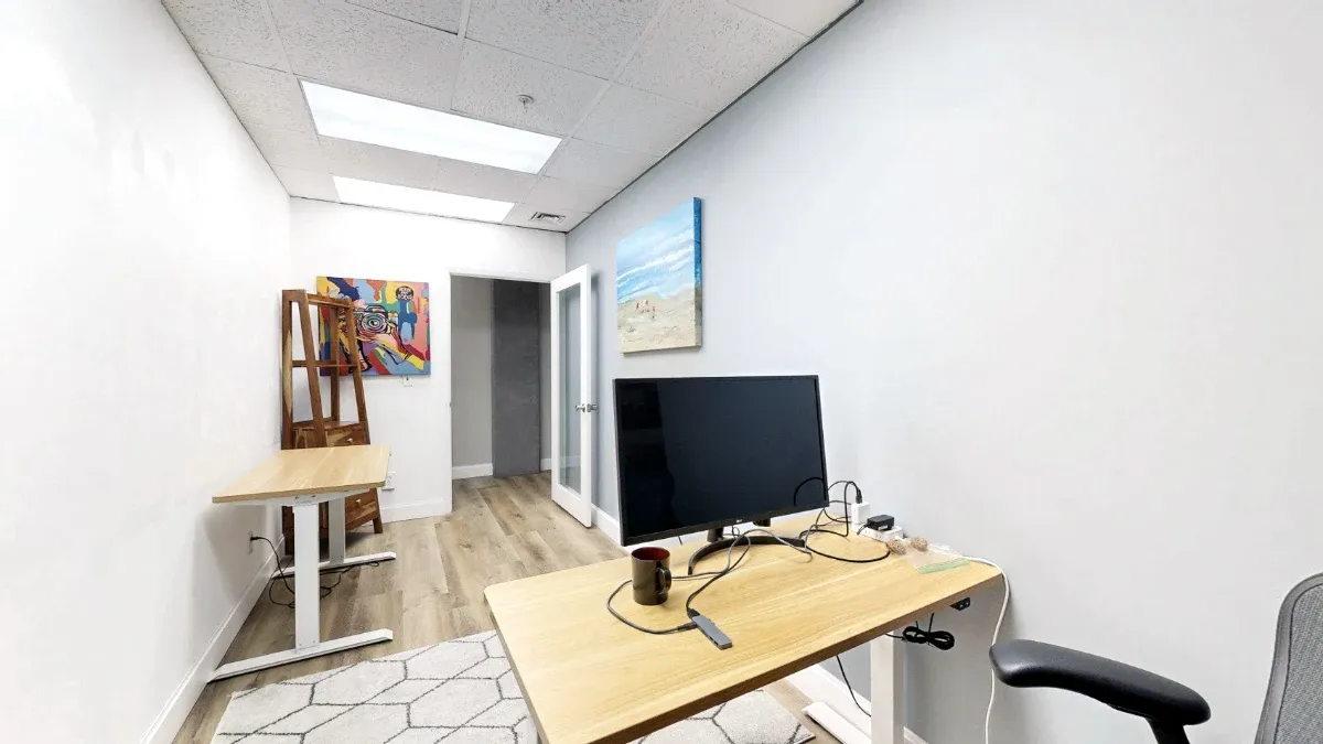 A monitor and mug sit on the foreground desk, with a second desk, colorful artwork and an open glass-paneled door beyond.