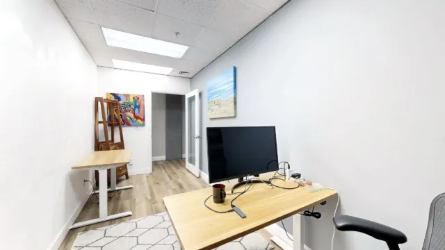 A monitor and mug sit on the foreground desk, with a second desk, colorful artwork and an open glass-paneled door beyond.
