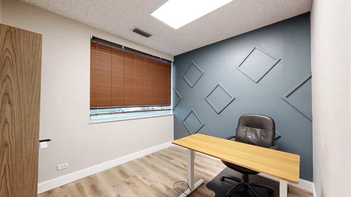 Office 207 with a wood-top desk, black chair, shaded window and blue-gray wall with geometric trim.