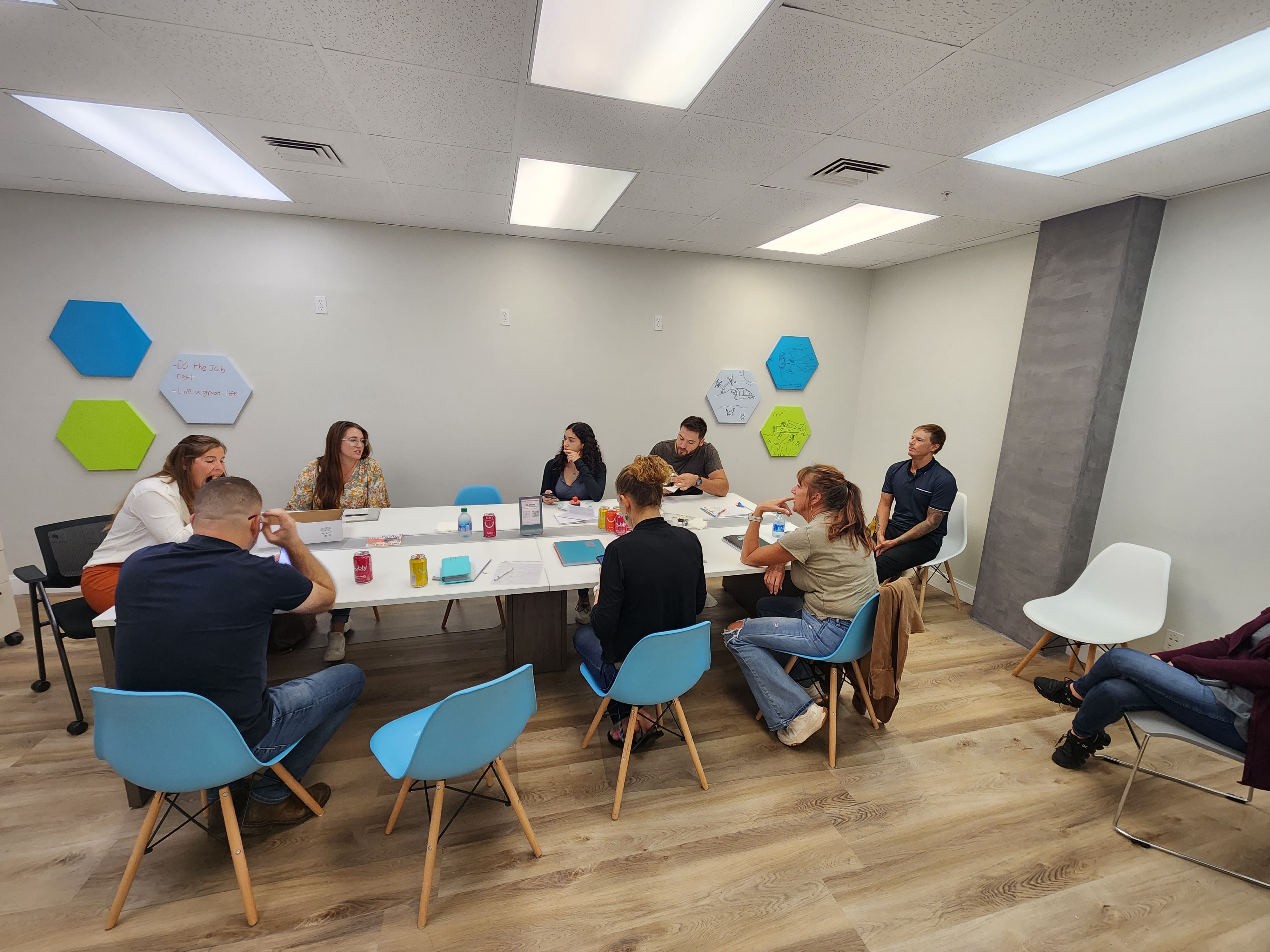 People sit around white tables with notebooks and drinks in a meeting room with colorful hexagonal wall panels.