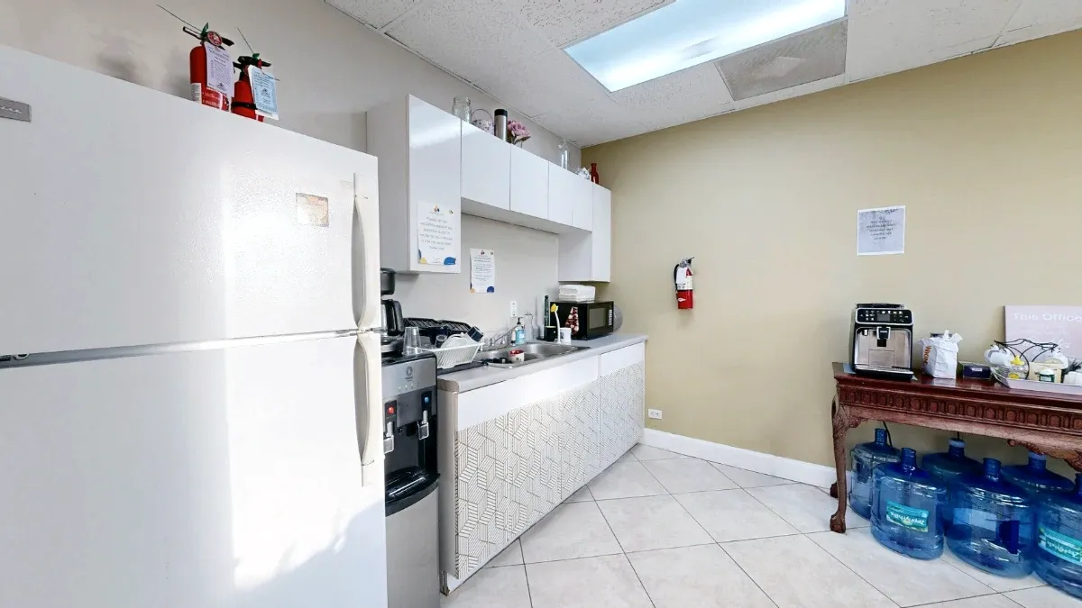 A white refrigerator stands beside the kitchenette sink and cabinets, with a coffee station and water jugs to the right.