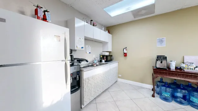 A white refrigerator stands beside the kitchenette sink and cabinets, with a coffee station and water jugs to the right.