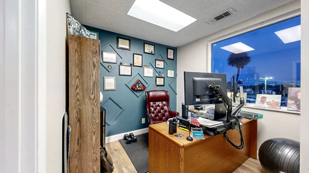 Office 212 with a wooden desk, burgundy chair, framed displays on a teal wall and a large window overlooking A1A.
