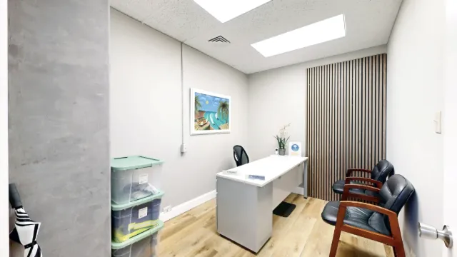 Doorway view of a white desk, black chairs, wooden slat wall decor and framed beach artwork in a windowless office.