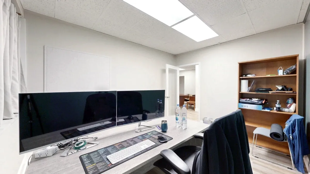 Small office with two large monitors on a desk, an office chair, wooden shelving, and an open door into the suite.