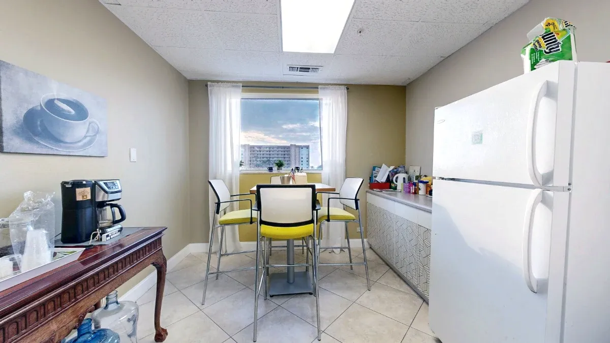 A tall lunch table and yellow-cushioned chairs sit by a curtained window between the coffee station and a refrigerator.