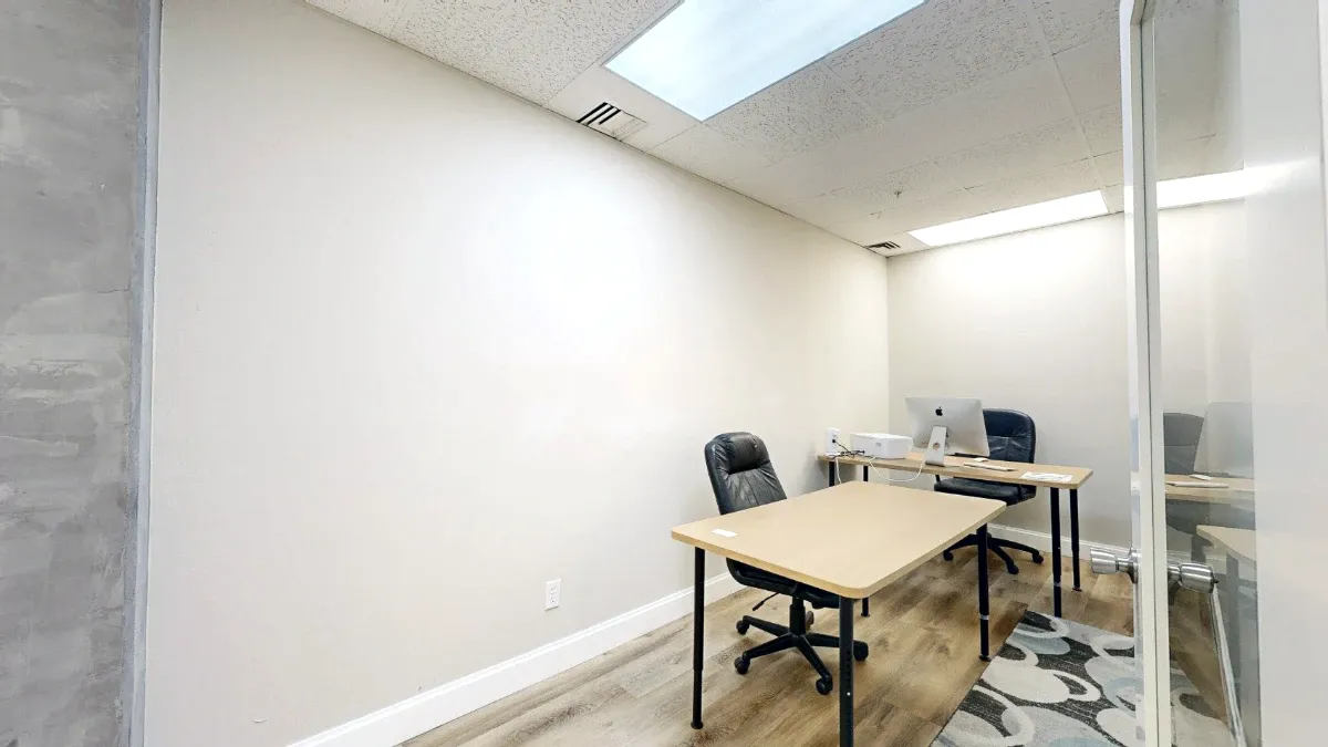 Two light wood desks with black rolling chairs line the narrow office beside an open glass-paneled door.