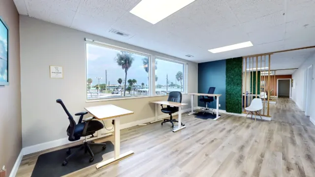 Daylight falls across three window-side desks, with a white accent chair beside a green panel and wooden slats.