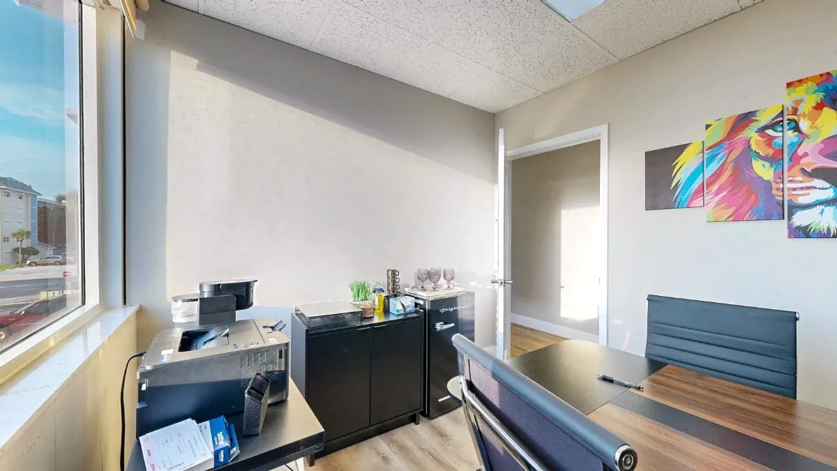 Office 201 with a wood-tone desk, black chairs, a mini fridge, glassware and colorful lion artwork.